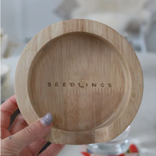 Load image into Gallery viewer, A hand holding a round, light-colored wooden tray for coasters with the brand name 'SEEDLINGS' debossed in the center. The background is a soft, out-of-focus indoor setting.