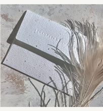 Load image into Gallery viewer, Envelope with the word 'PLANTABLE ENVELOPE' debossed, arranged diagonally on a light stone-like surface with dried beige feathers casting shadows.