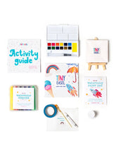 Load image into Gallery viewer, A flat-lay arrangement of a children's art activity kit on a white background, including an activity guide, watercolor paint palette, watercolor crayons, watercolor paper pad, paintbrush, tape, and a small easel card.