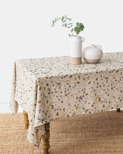 Load image into Gallery viewer, magic linen tablecloth in pressed botanical flower print on a natural linen background, draped over a long table with spindle legs on a jute rug. on top of the table are two stoneware vessels, one with a sprig of eucalyptus