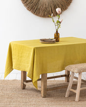 Load image into Gallery viewer, magic linen tablecloth in mossy sulfur yellow draped over a rustic wood table set with a tack of plates and a floral arrangement. a bench sits next to the table and a woven jute rug is beneath