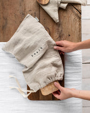 Load image into Gallery viewer, overhead shot of kitchen scene showing two hands pulling out a loaf of bread from a neutral linen drawstring bread bag, stamped with the word 'bread' on a wooden cutting board