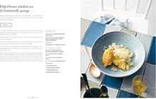Load image into Gallery viewer, A magazine-style spread featuring a recipe article titled 'Elderflower sherbet ice &amp; buttermilk sponge' on the left and a plated food photograph on the right. The food photo shows a golden sponge cake with ice on a blue plate over a tiled surface, with a spoon, glass, and kitchen setting visible.
