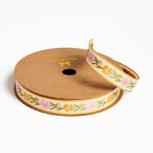 Load image into Gallery viewer, A spool of decorative ribbon on a plain white background. The ribbon has a white base with a repeating floral pattern in pink, yellow, and green, bordered by a gold edge.

