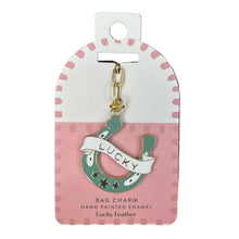 Load image into Gallery viewer, A hand-painted enamel bag charm shaped like a green horseshoe with a white banner reading 'LUCKY' and small feather details, attached to a pink and white display card. 
