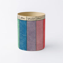 Load image into Gallery viewer, A cylindrical decorative container or candle holder with a vintage color-blocked design featuring vertical stripes in green, blue, violet, mauve, and red, with a beige band at the top labeled in French.

