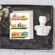 Load image into Gallery viewer, A greeting card with a whimsical tea-themed illustration sits on a dark textured tray, styled with dried flowers and a white ceramic bust.
