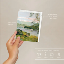 Load image into Gallery viewer, A hand holds a seed paper envelope and greeting ard featuring a landscape illustration, with text callouts pointing to foil details and plantable seed paper envelope features on a neutral background.
