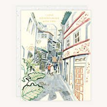 Load image into Gallery viewer, A greeting card featuring a watercolor illustration of a narrow European street with the text 'I LOVE ADVENTURING WITH YOU' at the top, shown with a matching envelope behind it on a white background.
