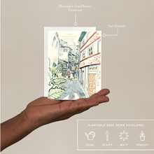 Load image into Gallery viewer, A hand holds a greeting card and seed paper envelope featuring a watercolor illustration of a narrow street with buildings and green plants. White callout lines point to the envelope and label its features, including 'Plantable Seed Paper Envelope' and 'Foil Details.
