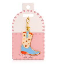 Load image into Gallery viewer, A hand-painted enamel keychain charm shaped like a cowboy boot, displayed on its pink retail card. The boot is cream-colored with orange star patterns, light blue sides, and orange soles, hanging from a gold-tone clasp.
