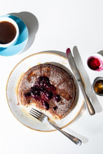 Load image into Gallery viewer, A top-down view of a plate of pancakes topped with powdered sugar and blueberry syrup, served with a fork and knife on a white surface, alongside a cup of coffee and small bowls of syrup.
