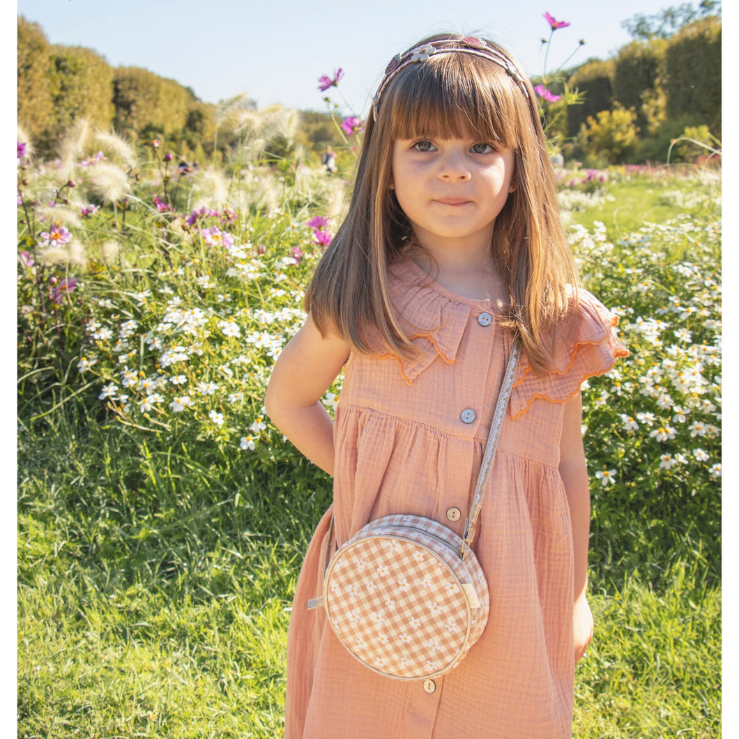 little girl standing in a flower field wearing a peach dress and rock-a-hula's cross-body circle bag in a neutral gingham pattern that has embroidered daisies all over it and a top zipper