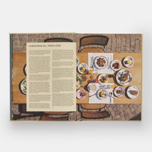 Load image into Gallery viewer, An open book spread showing a printed article titled 'CHRISTMAS ALL YEAR LONG' with a large text panel on the left and a color photograph of a dining table set with plates and food on the right, photographed against a light gray background.
