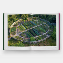 Load image into Gallery viewer, An open hardcover book on a light gray background, displaying a large aerial photograph of a terraced vegetable garden on the left page.
